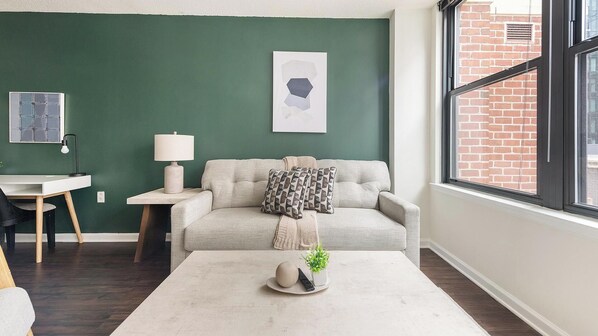 Uma sala de estar com parede verde, um sofá, uma mesa de centro, uma luminária e uma janela com vista para uma parede de tijolos.