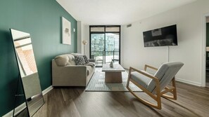 A modern living room with a sofa, a wooden rocking chair, a coffee table, and a TV on the wall. - Landing | Stunning 2BD, Courtyard, Gym (Washington)