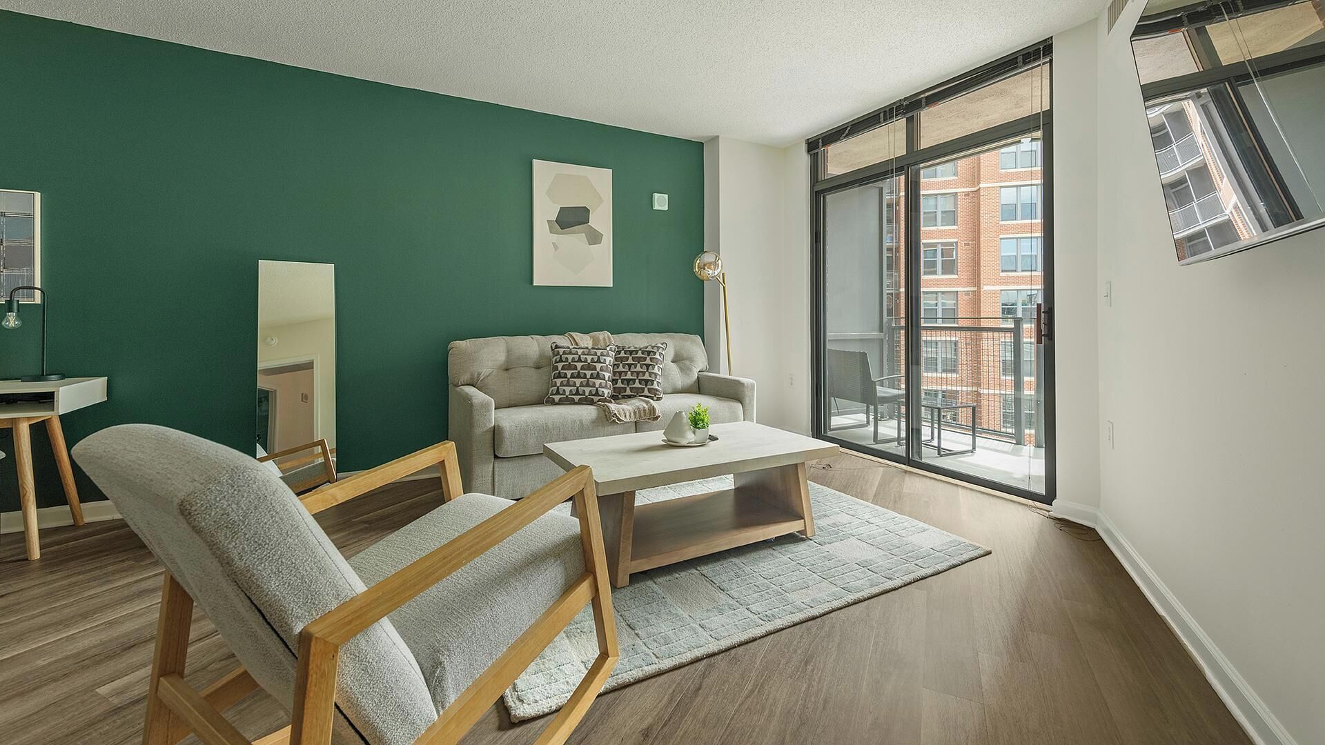 Uma sala de estar moderna com uma parede verde de destaque, um sofá, uma mesa de centro e uma janela grande com vista para edifícios.