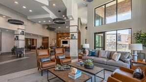 A spacious lobby with leather armchairs, a sofa, and a coffee table. There are pendant lights, a fireplace, and large windows offering views of the outside. - Landing | Stylish 1BD, Gym, Game Room (Fort Worth)