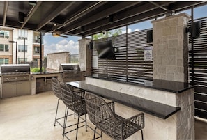 An outdoor kitchen with a stone wall, a grill, and two wicker bar stools. - Landing | Chic 1BD, Gym, Game Room (Fort Worth)