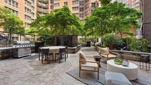 Eine Terrasse mit Grill, Esstisch und Stühlen, umgeben von Grünpflanzen und einer Ziegelmauer.