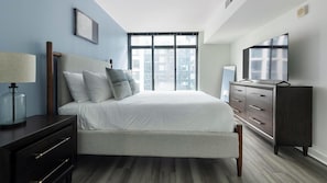 Ein modernes Schlafzimmer mit Bett, Kommode, Fernseher und Fenster mit Blick auf die Stadt.