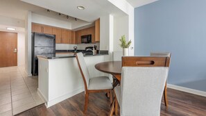 A compact kitchen with wooden cabinets, a refrigerator, and a dining area with a table and chairs. - Landing | Stunning 2BD, Courtyard (Washington)
