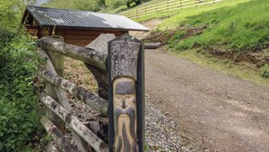 Una valla de madera rústica con una figura de madera tallada, un sendero de tierra y una estructura de madera con techo de metal en un paisaje verde exuberante.