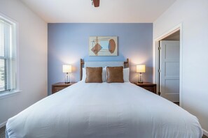 Een hotelkamer met een groot bed, twee nachtkastjes met lampen, een kunstwerk aan de muur, en een deur aan de rechterkant.