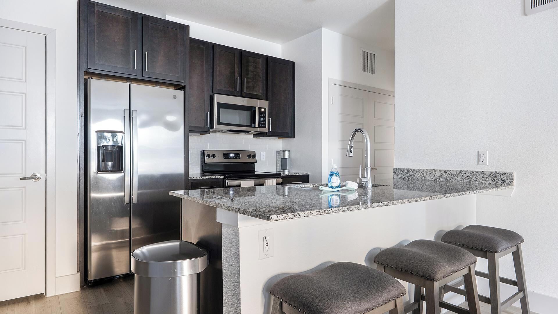 A modern kitchen with dark cabinets, stainless steel appliances, a granite countertop, and bar stools.