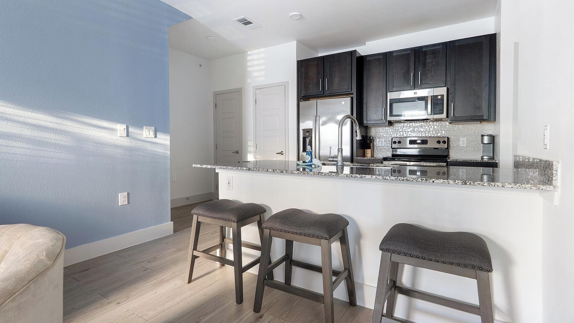 A modern kitchen with dark cabinets, a stainless steel refrigerator, and a microwave. There are three bar stools and a white countertop.