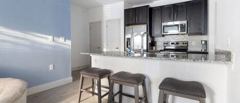A modern kitchen with dark cabinets, a stainless steel refrigerator, and a microwave. There are three bar stools and a white countertop.