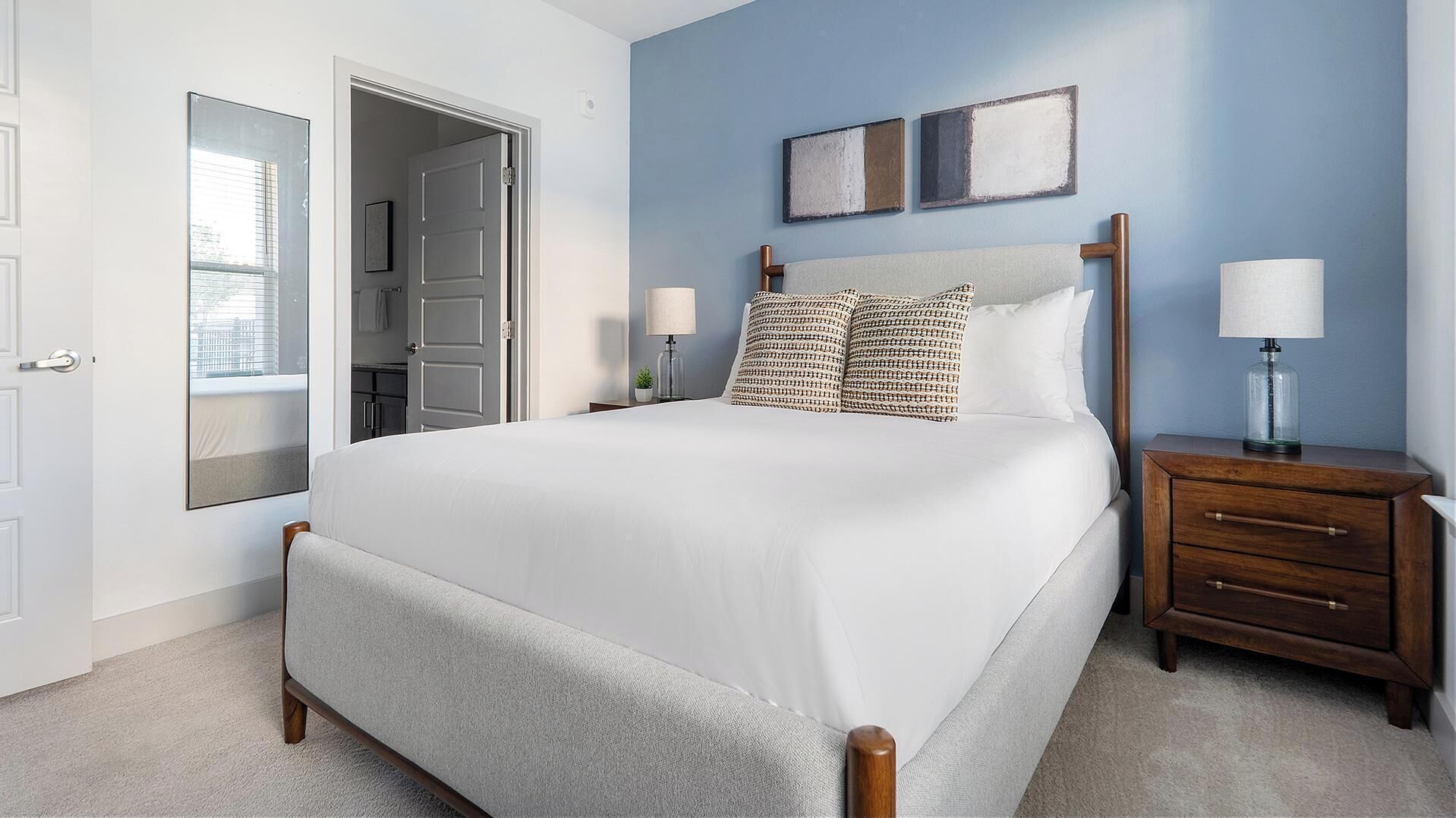 A bedroom with a bed, bedside tables, a nightstand, a mirror, and a door leading to a bathroom.