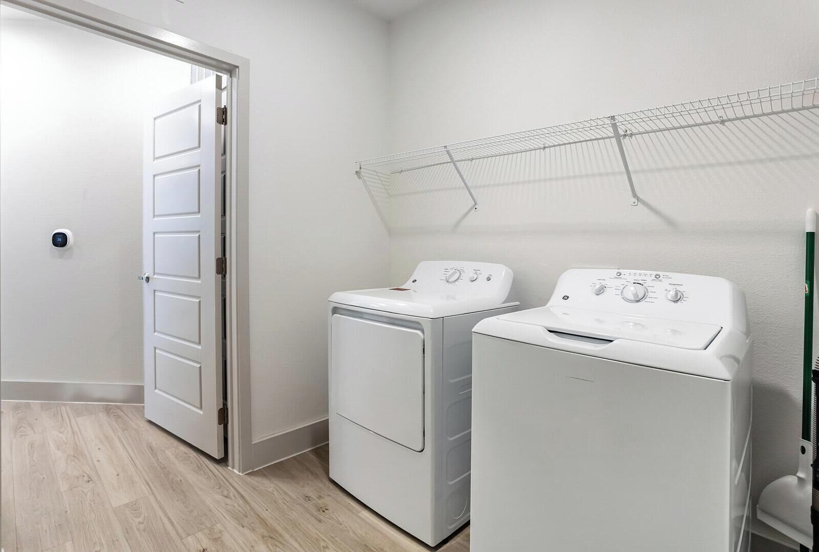 A laundry room with two white front-loading washers and dryers, a metal drying rack, and a door leading to another room.