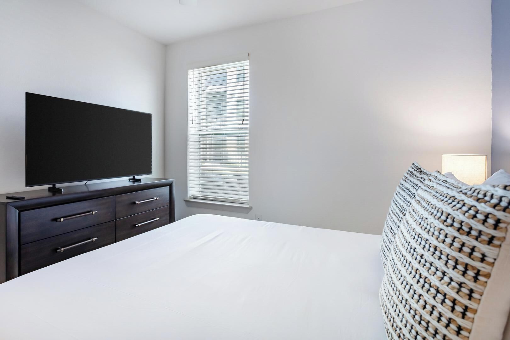 Een slaapkamer met een bed, een kaptafel, een televisie en een raam met jaloezieën.