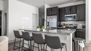 A modern kitchen with dark cabinets, stainless steel appliances, a granite island with bar stools, and a trash can. - Landing | Stylish 2BD, Gym, Pool (Austin)