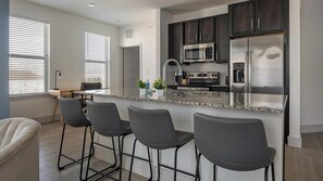 A modern kitchen with dark cabinets, a stainless steel refrigerator, a microwave, and a granite countertop. There are four bar stools and a small dining table with chairs. - Landing | Spectacular 2BD, Gym, Pool (Austin)