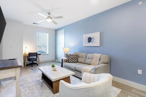A modern living room with a ceiling fan, a sofa, a coffee table, a desk with a chair, and a window with blinds. - Landing | Stylish 1BD, Gym, Pool (Austin)