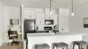 A modern kitchen with white cabinetry, a stainless steel refrigerator, a microwave, and a large island with bar stools. - Landing | Stylish 1BD, Gym, Clubhouse (Asheville)