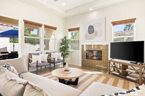 A modern living room with a sofa, coffee table, TV, fireplace, and large windows. - Desert Jewel | Puerta Azul Retreat Near PGA West (La Quinta)