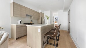 A modern kitchen with wooden cabinets, a central island, bar stools, and a visible dining area. - Landing | Incredible 2BD in Logan Circle (Washington)