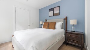 A neatly made bed with a white duvet, two brown pillows, a wooden headboard, and a nightstand with a lamp and a glass bottle. - Landing | Amazing 1BD in Logan Circle (Washington)