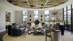 Uma sala de estar moderna com uma mesa de centro, dois sofás e uma poltrona. Janelas amplas proporcionam iluminação natural e vista para o exterior.