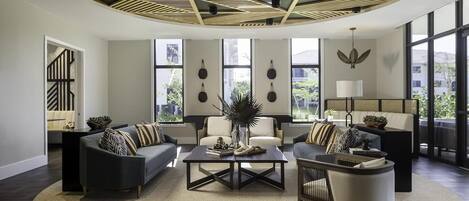 A modern living room with a central coffee table, two sofas, and a chair. There are large windows providing natural light and views of the outdoors.