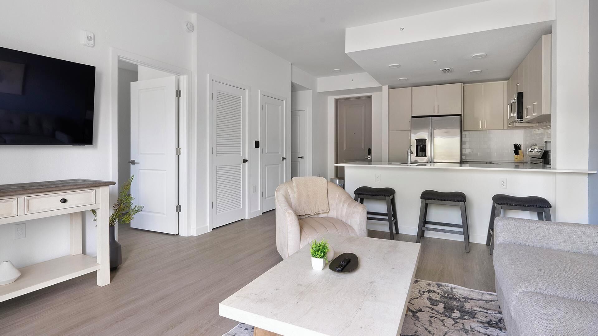 A modern living room with a sofa, a coffee table, and a TV. There is a kitchen area with a refrigerator and a sink.