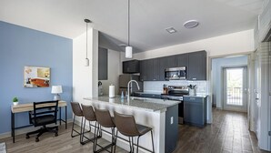 Una cocina moderna con una isla central, taburetes de bar, una zona de comedor con escritorio y sillas, y vistas al exterior a través de una puerta.