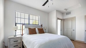 Un dormitorio con cama, mesitas de noche, una ventana con persianas, una puerta y un ventilador de techo.