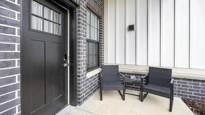 Eine schwarze Tür mit Fenster darüber, eine Ziegelwand und zwei Korbstühle davor.