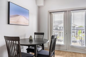 A dining area with a round table and four chairs, a vase on the table, and a large window with blinds. - Landing | Amazing 1BD, Gym, Clubhouse (McCordsville)