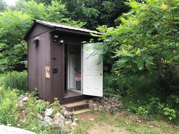 Un petit abri en bois avec une porte blanche, entouré d’une végétation luxuriante et de rochers.