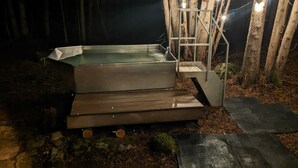 Een metalen jacuzzi met metalen trapje en treden, omringd door bomen en verlicht met lichtslingers.