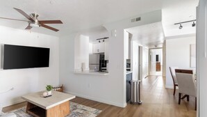 Una sala de estar moderna con un televisor de pantalla plana, un ventilador de techo, una zona de comedor con mesa y sillas, y una cocina visible a través de un vano abierto.