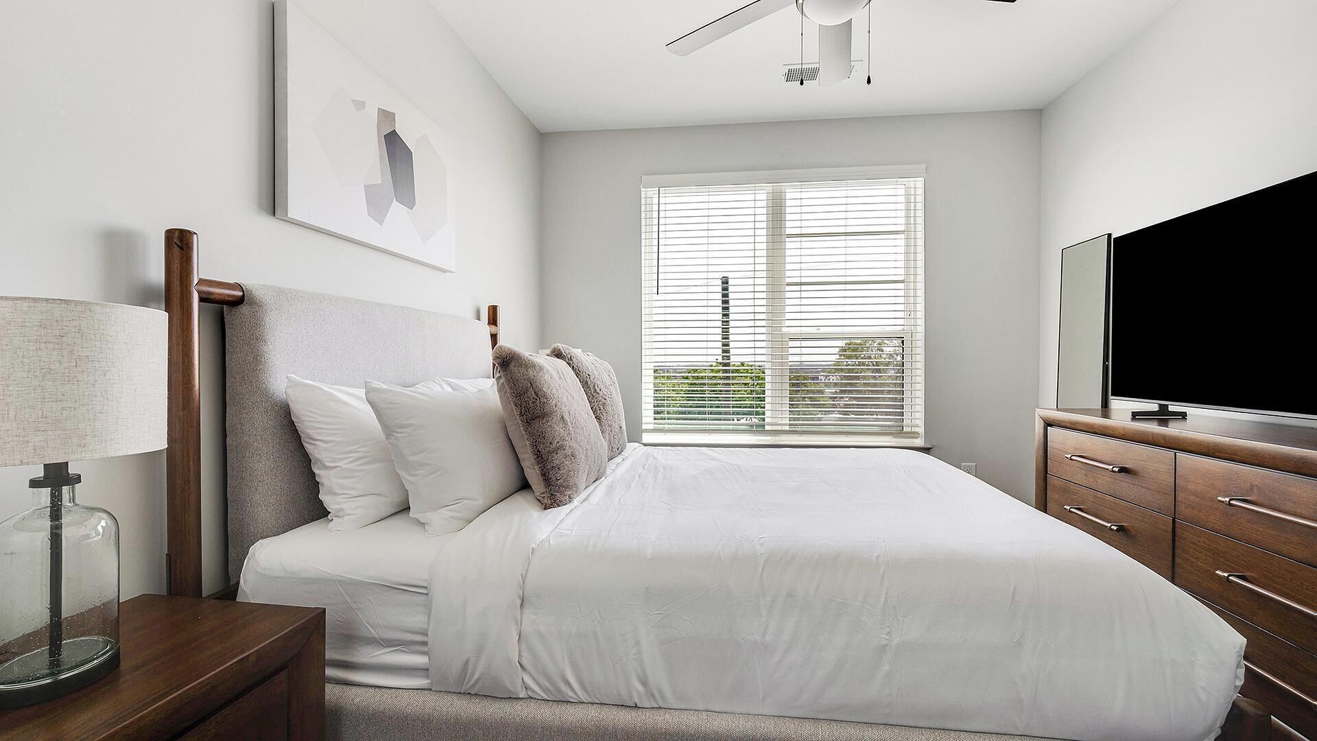 A bedroom with a bed, a TV, a nightstand, a lamp, and a window with blinds.