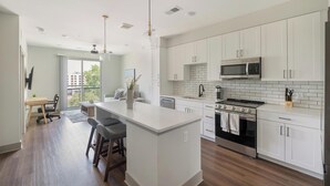 A modern kitchen with white cabinetry, a central island, stainless steel appliances, and a dining area with a view. - Landing | Chic 1BD, Courtyard, Gym (Nashville)