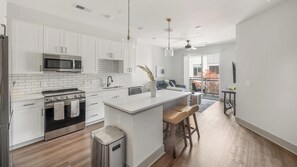 A modern kitchen with white cabinetry, a stainless steel stove, and a large island with bar stools. - Landing | Stylish 1BD, Courtyard, Yoga Studio (Nashville)