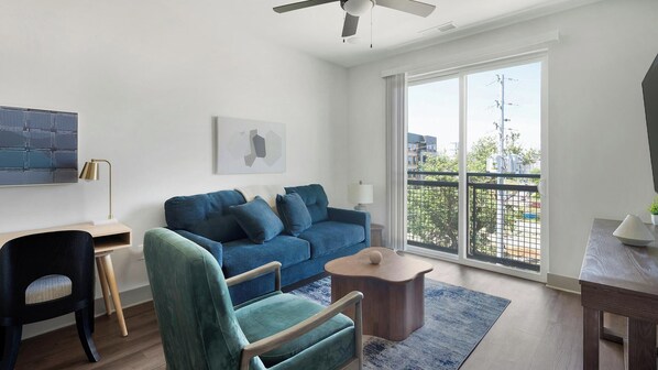 A modern living room with a blue sofa, a wooden chair, a small table, and a large window with a view of buildings and trees. - Landing | Stylish 1BD, Gym, Courtyard (Nashville)