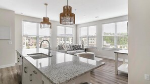 A modern kitchen with a large island, pendant lights, and a view of buildings through large windows. - Landing | Beautiful 2BD, Clubhouse, Pool (Conroe)