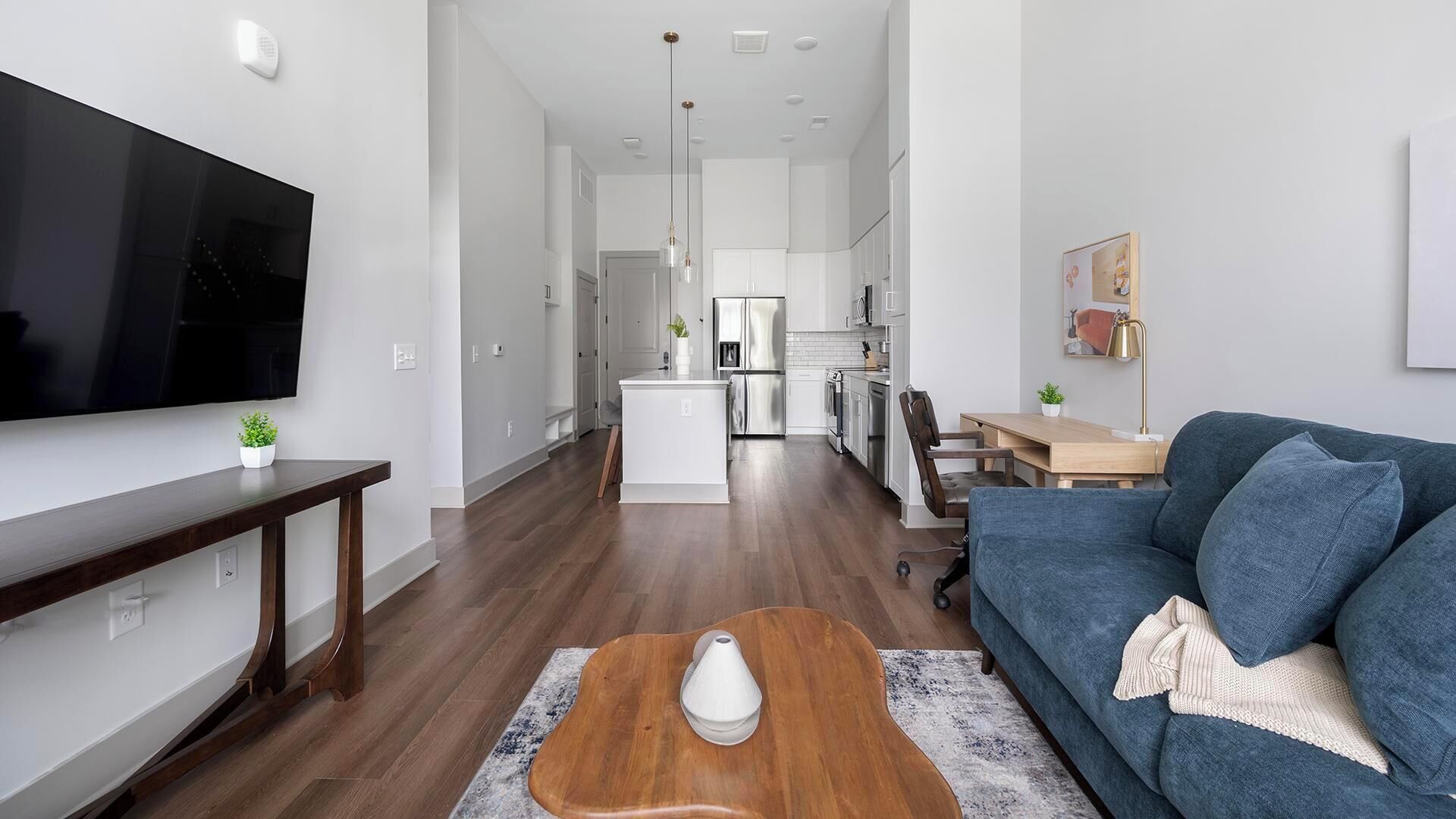 Una sala de estar moderna con un televisor de pantalla plana, un sofá azul, una mesa de centro de madera y una cocina al fondo.