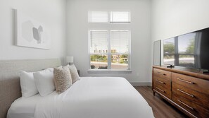A bedroom with a bed, a dresser, a television, and a window with blinds. - Landing | Modern 1BD, Gym, Courtyard (Nashville)