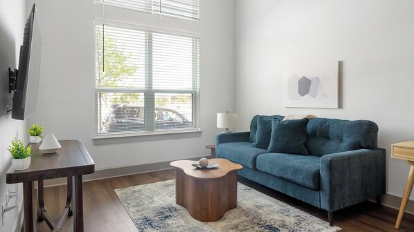 A living room with a blue sofa, a wooden coffee table, a TV mounted on the wall, and a window with blinds. - Landing | Modern 1BD, Gym, Courtyard (Nashville)
