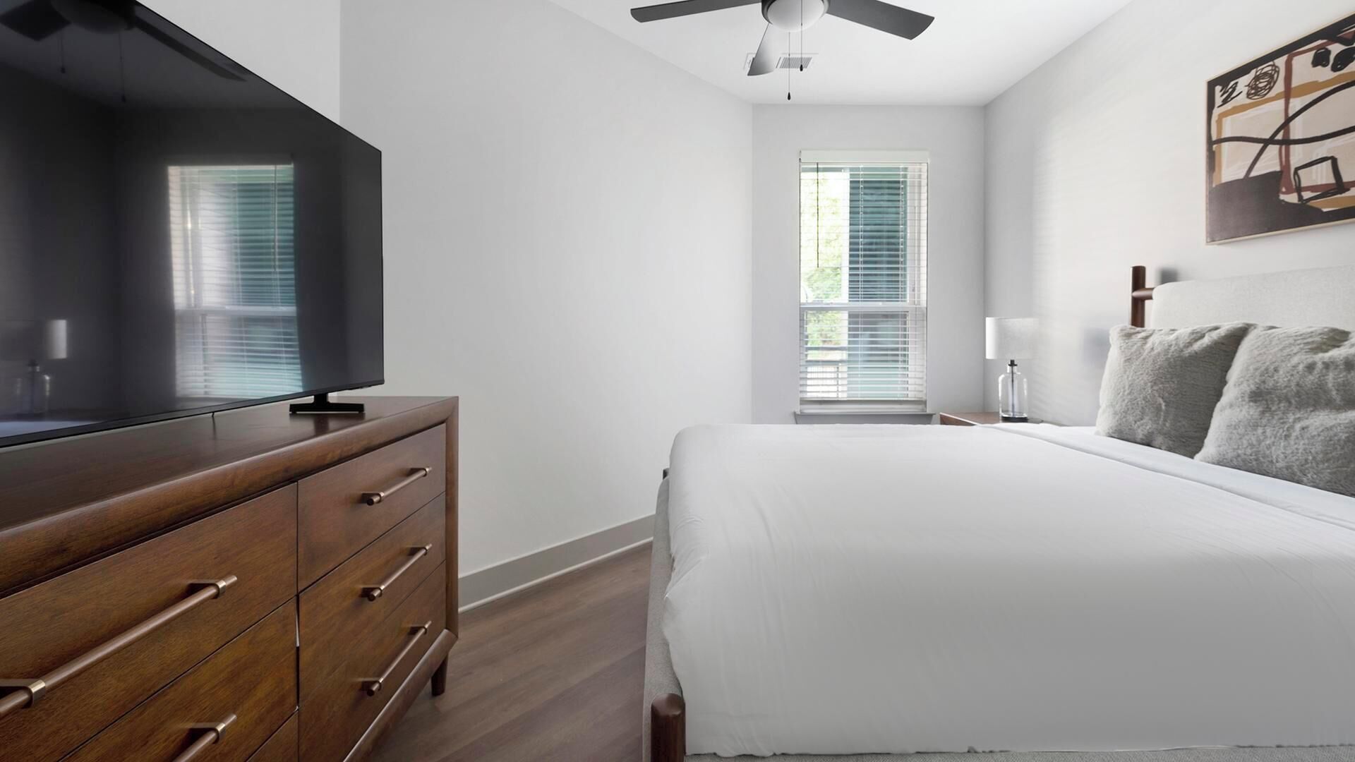 Ein Schlafzimmer mit Fernseher auf einer Holzkommode, einem Bett mit weißer Bettwäsche, einem Deckenventilator und einem Fenster mit Jalousien.