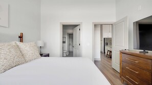 A bedroom with a bed, bedside table, lamp, dresser, TV, and a view into the kitchen and bathroom. - Landing | Chic 1BD, Courtyard, Gym (Nashville)