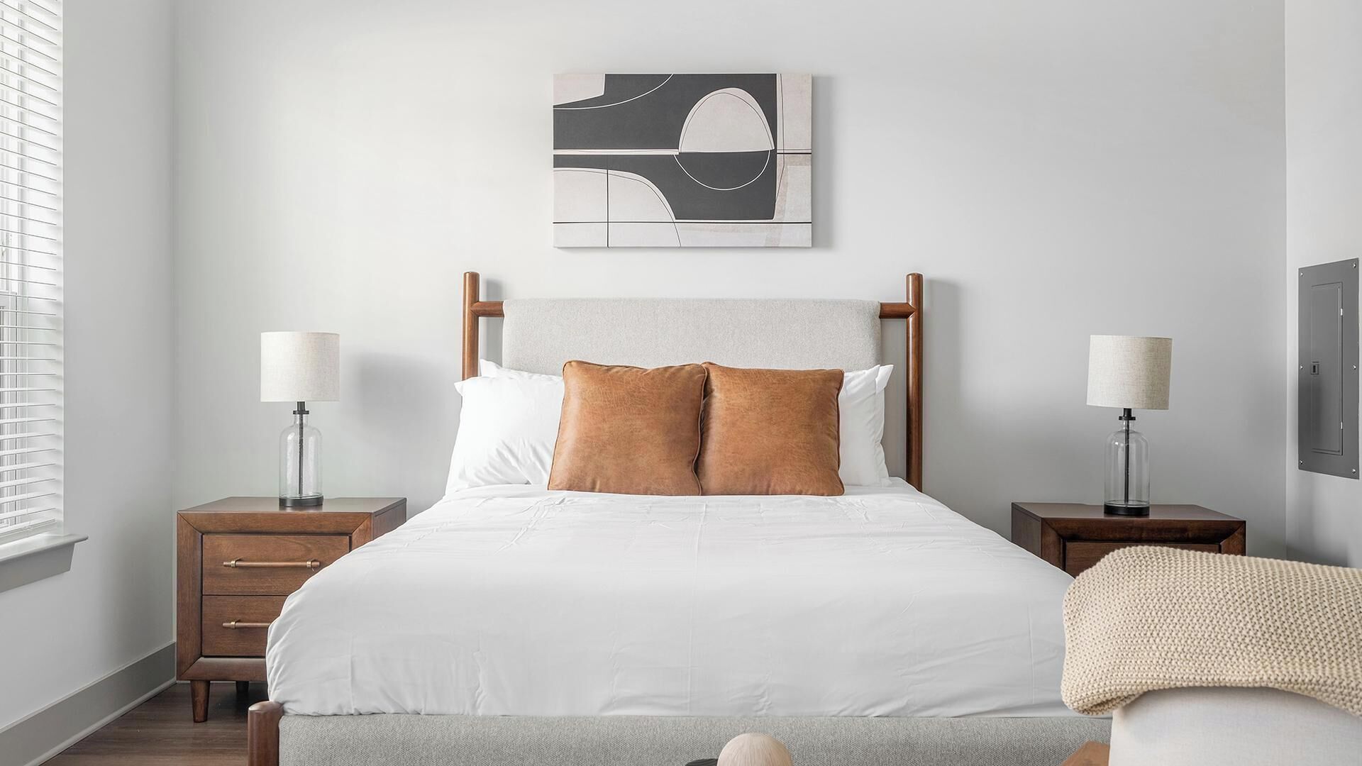 A bedroom with a bed, two bedside tables, a nightstand, a lamp, a blanket, and a wall art piece.