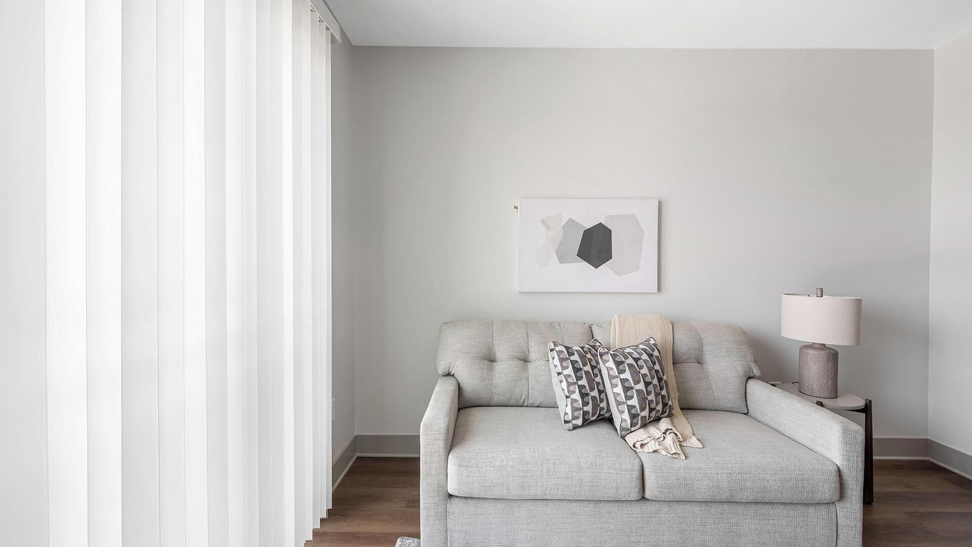 A neutral-toned living room with a grey sofa, a lamp, and a wall-mounted abstract art piece.