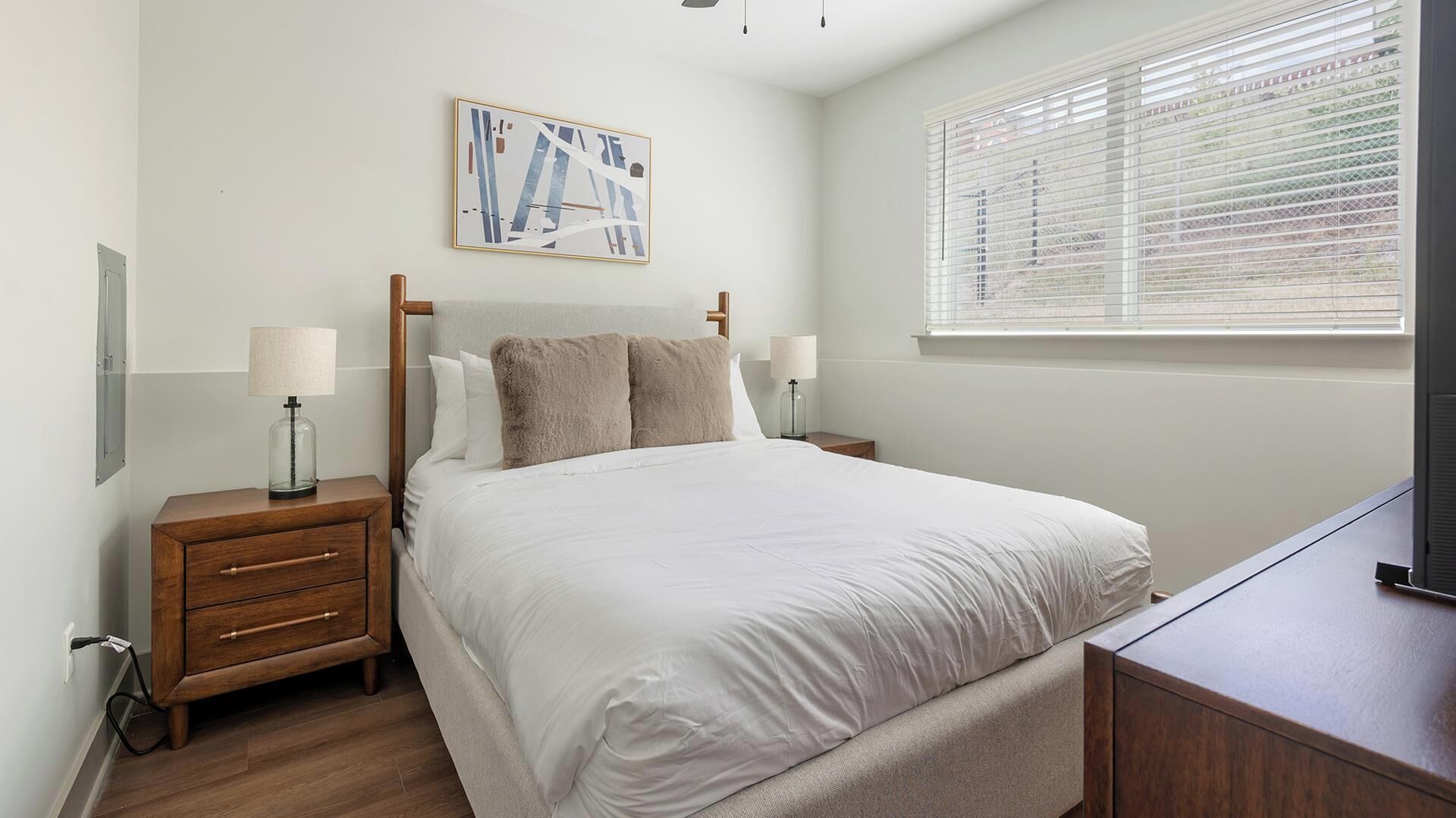 A bedroom with a bed, bedside tables, a lamp, a painting, and a window with blinds.