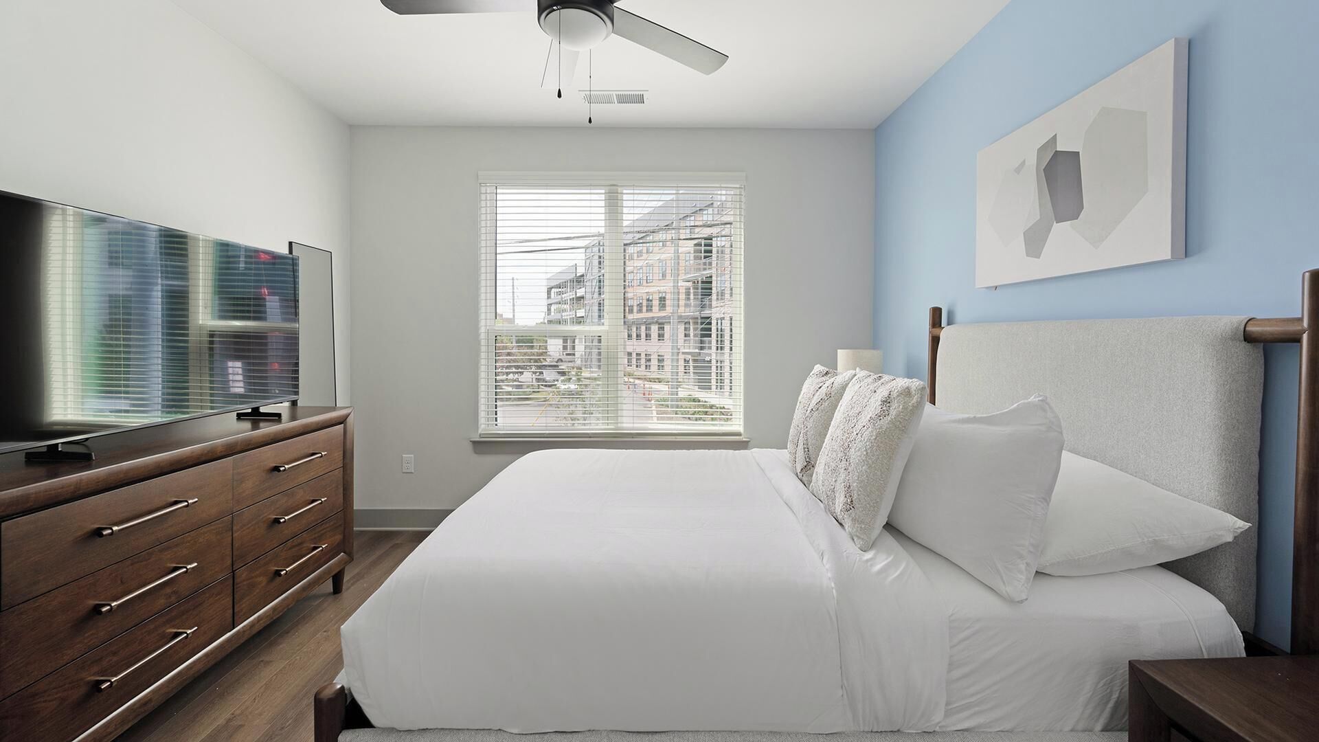 Ein Schlafzimmer mit einem Bett, einem Schminktisch, einem Fernseher, einem Fenster mit Jalousien und einem Deckenventilator.