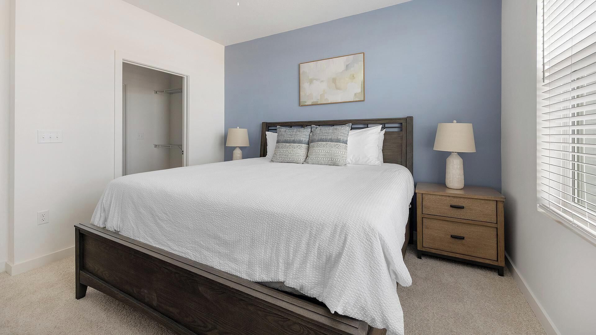 A bedroom with a bed, bedside tables, a nightstand, a door, and a window with blinds.