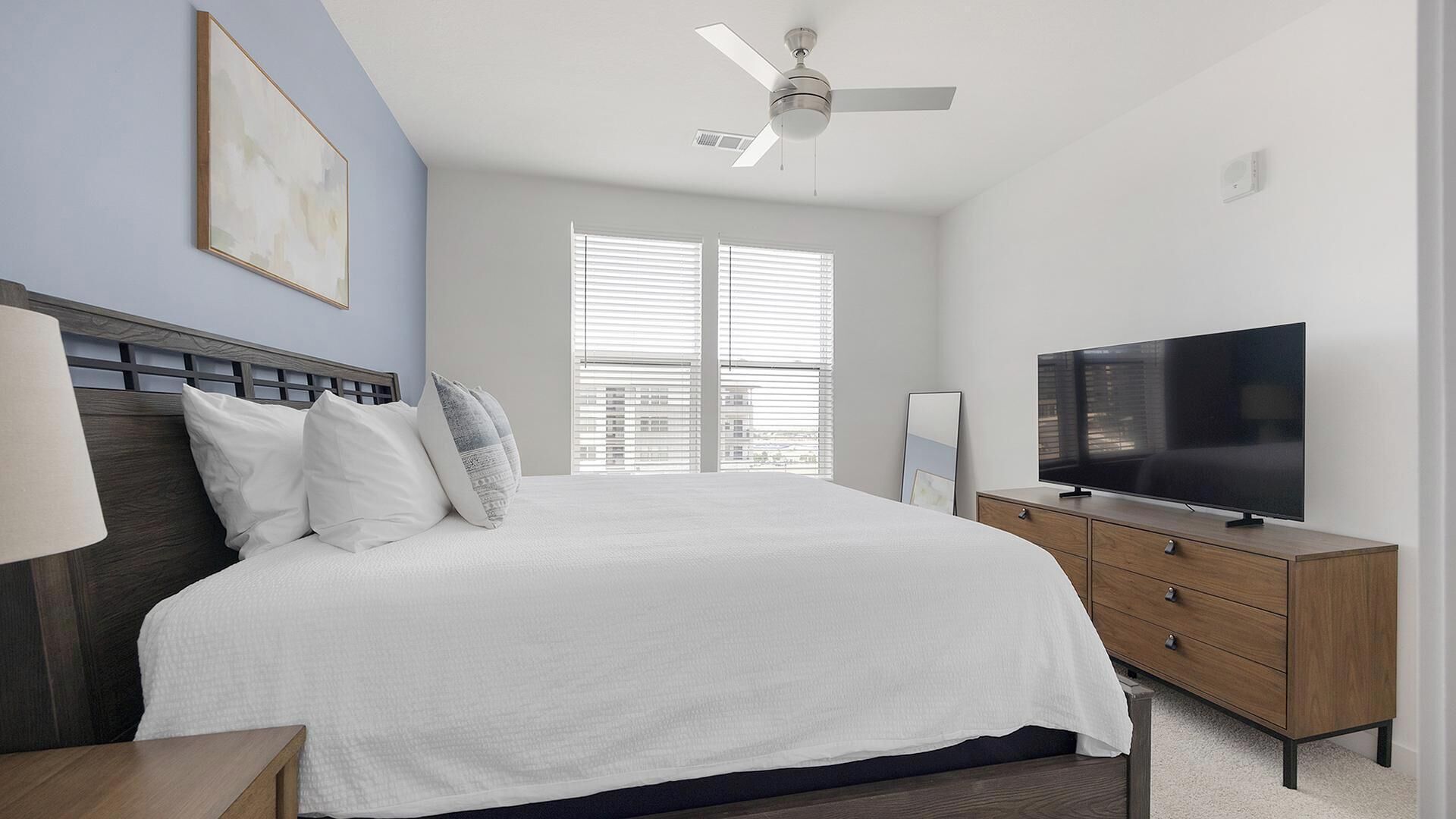 A bedroom with a bed, a TV, a ceiling fan, and a window with blinds.