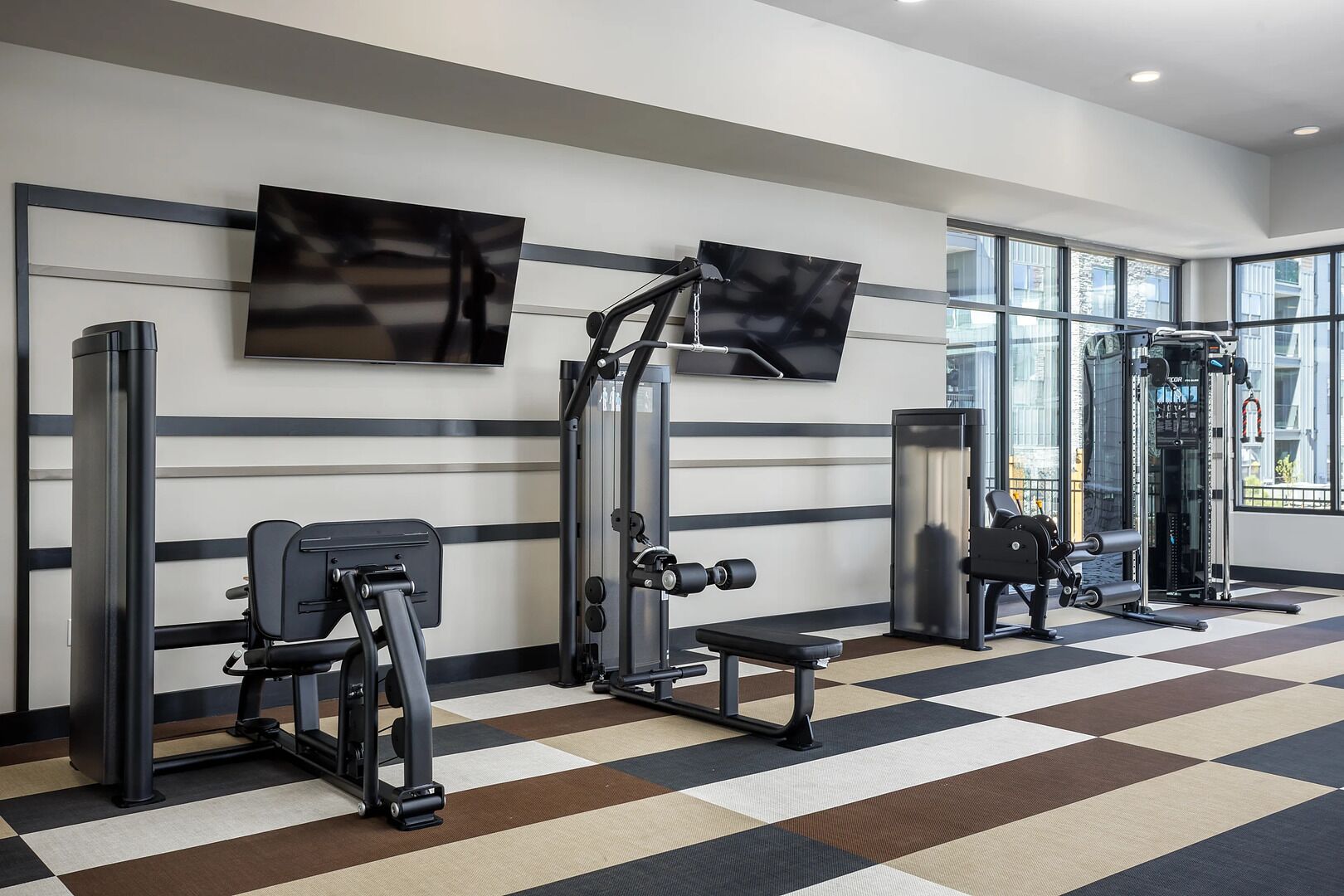 Een sportschool met trainingsapparatuur, grote ramen en een geruit vloertapijt.